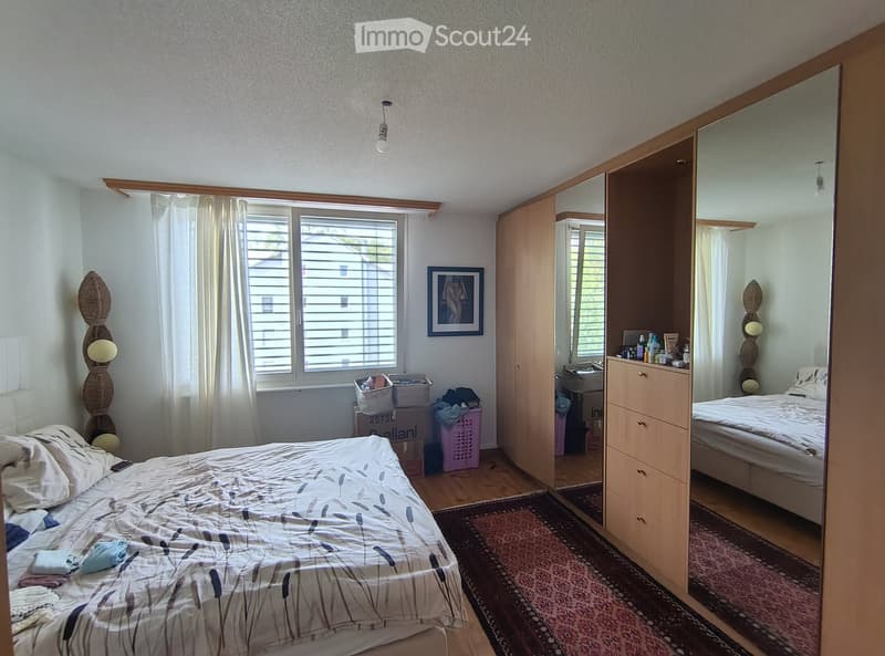 Well-lit bedroom with a large bed, wooden wardrobe, mirror, picture frame, and a small table with personal items.