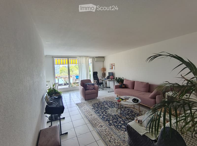 Modern living room with a pink sofa, armchair, coffee table, laptop, and plants. The room features large windows with blinds and a tiled floor.