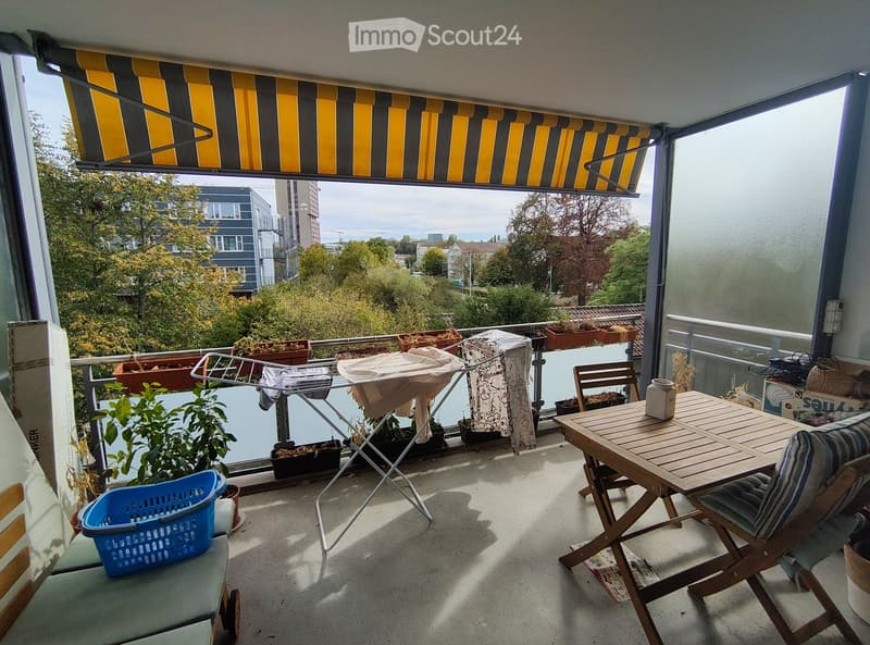 Balcony with a view of the city, featuring a wooden table, chairs, plants, a laundry drying rack, and a laundry basket.