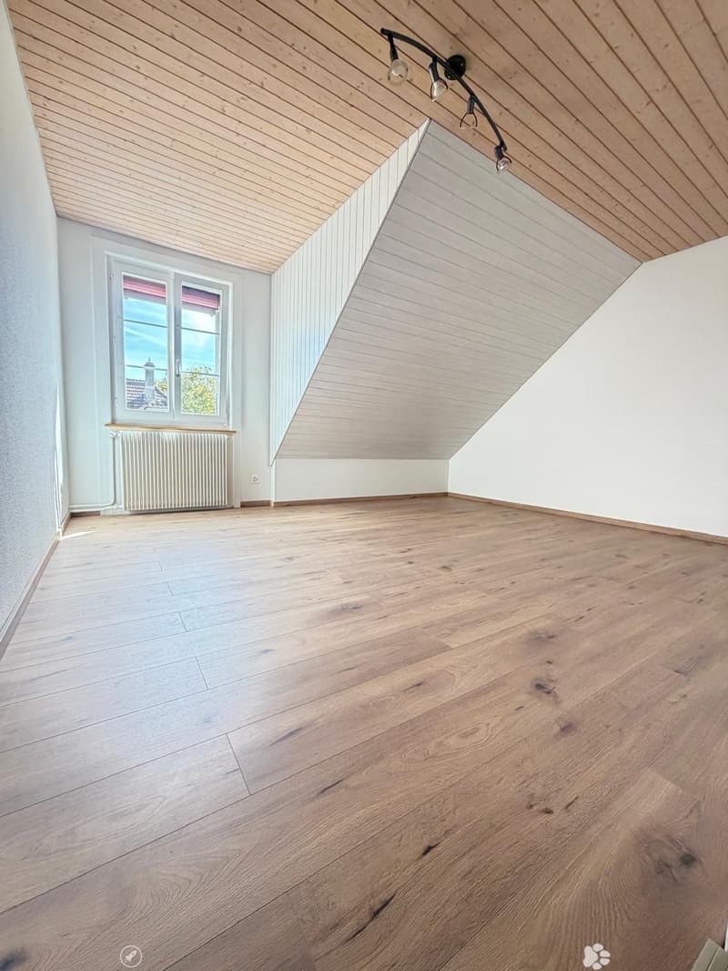 Empty attic room with wooden flooring, white walls, and a slanted wooden ceiling. Features a window with a view and a radiator below it.