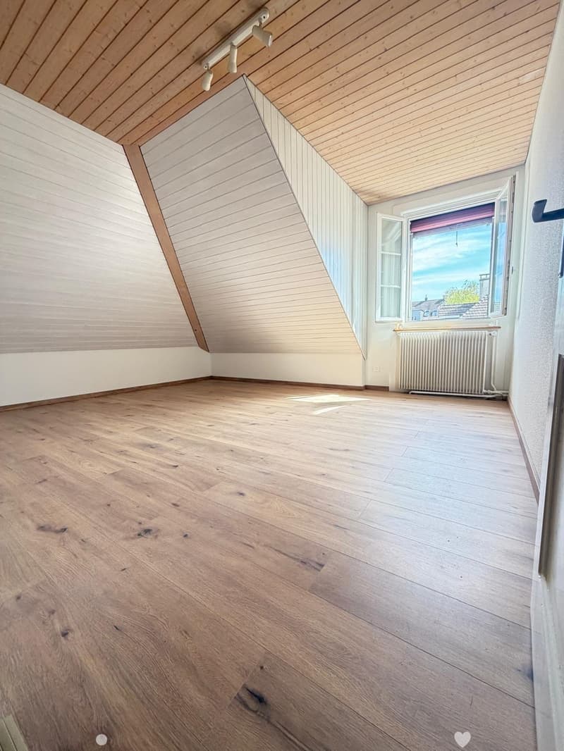 Empty attic room with wooden floors and slanted wooden ceilings, featuring large windows and a radiator.