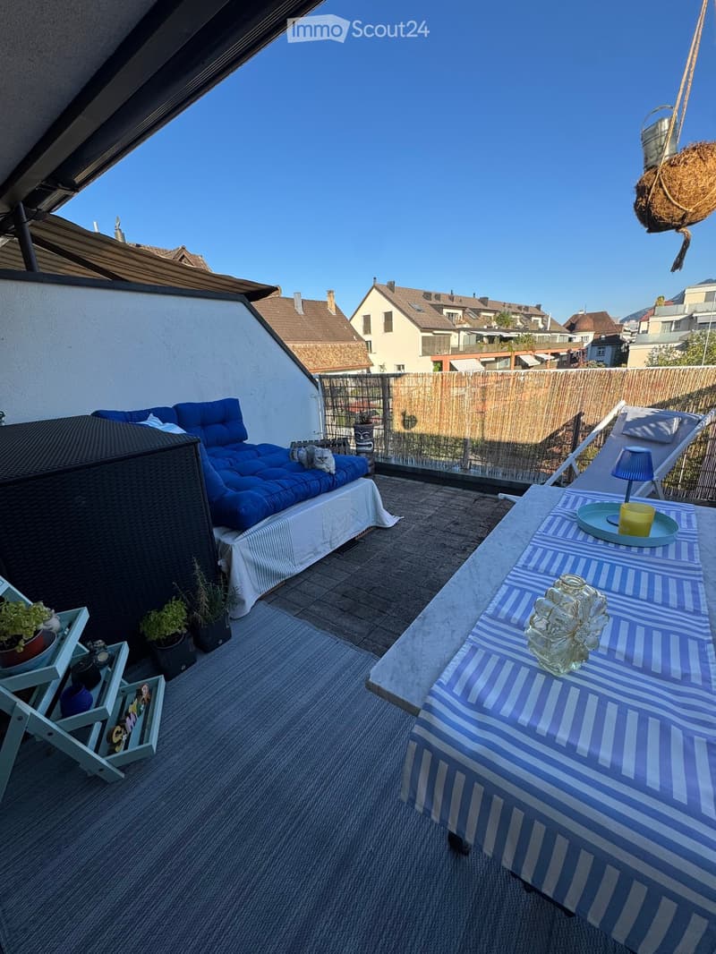 A furnished balcony with a blue sofa, a small table with a lamp, a hanging basket, potted plants, and a lounge chair.