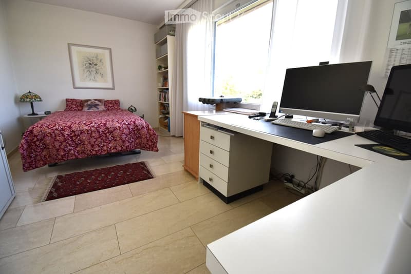 Bright bedroom with a large bed, a desk with a computer setup, a bookshelf, and a lamp. The room features large windows with white curtains and a tiled floor.