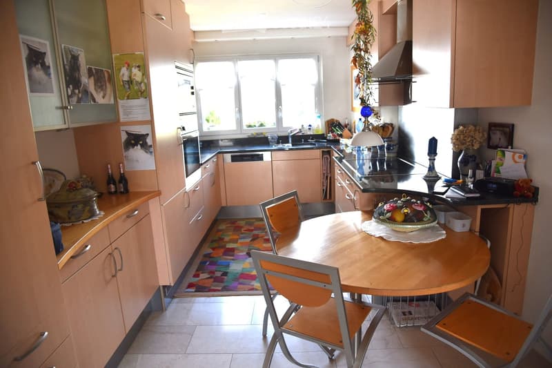 Bright kitchen with light wooden cabinets, a round wooden dining table with four chairs, a sink, and a stove. The room features a colorful rug, framed pictures, and various kitchen items on the counters.