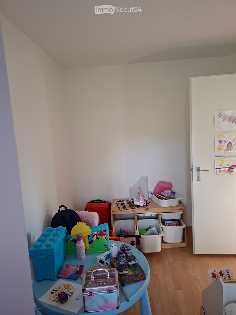 A small, cozy room with light-colored walls and wooden flooring. A blue table holds various items including a toy, a bottle, a cup, and shoes. A storage unit with drawers and shelves is positioned against the wall, containing toys and other items. A backp