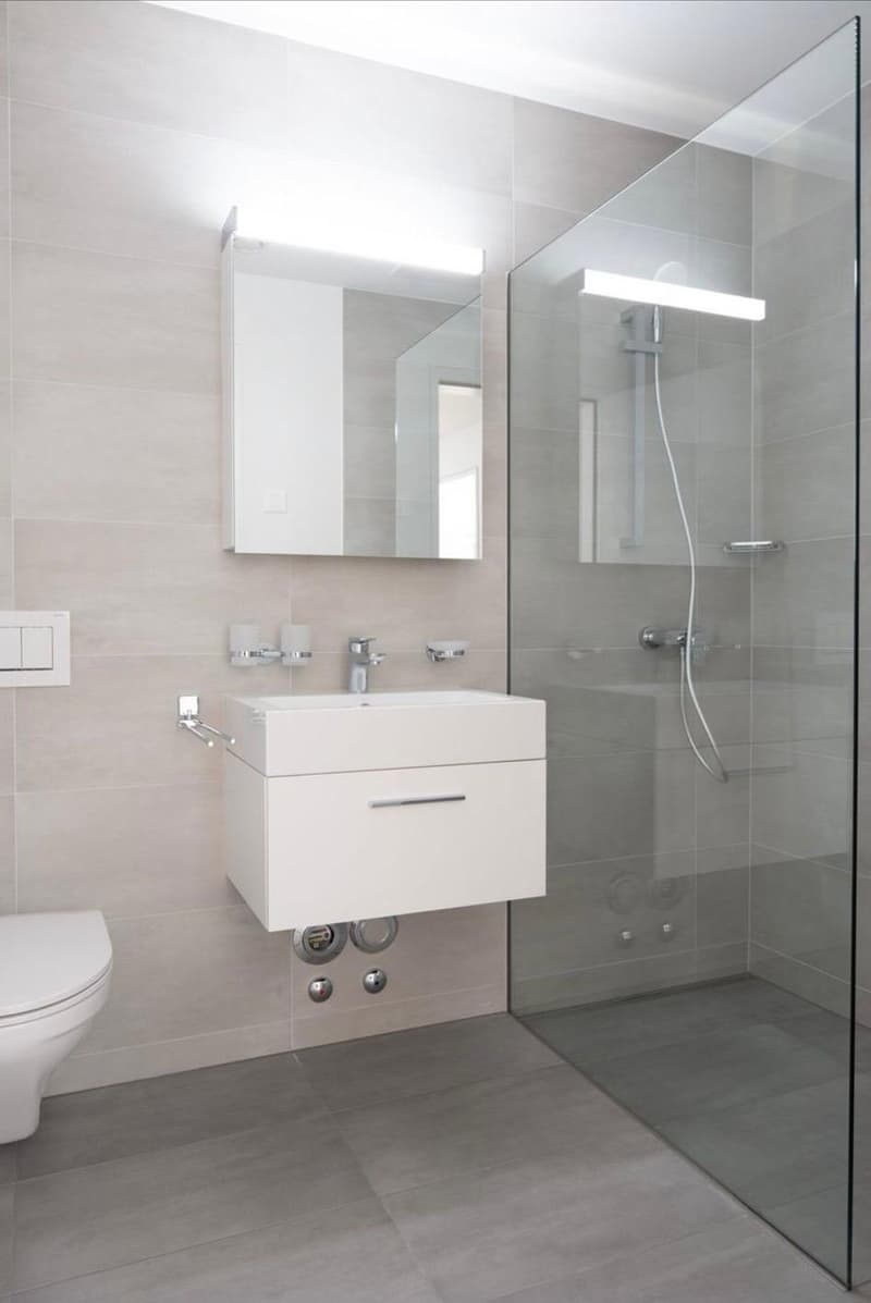 Modern bathroom with a white sink, mirror, toilet, and glass-enclosed shower. Light gray tiles and sleek fixtures.
