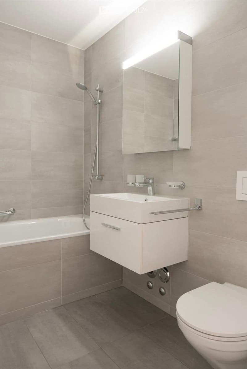 Modern bathroom with a white sink, mirror, toilet, bathtub, and showerhead. Light gray tiles on walls and floor.