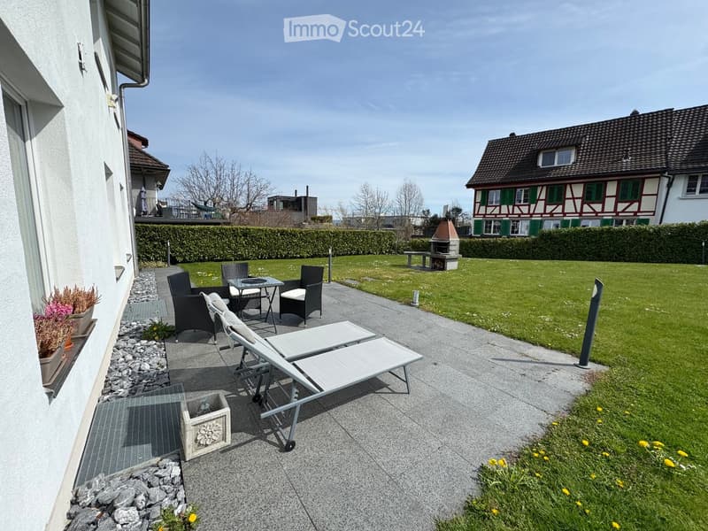 Garden area with a patio, lounge chairs, a small table, and a bench near a barbecue. The area is surrounded by a hedge and a lawn, with a house and additional outdoor furniture visible in the background.