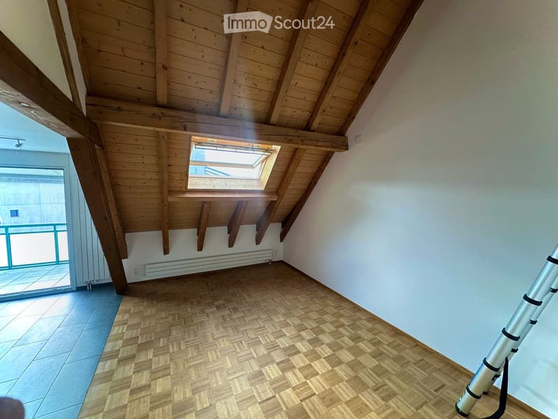 Empty attic room with wooden slanted ceiling, herringbone wooden floor, skylight window, and sliding glass door leading to a balcony.