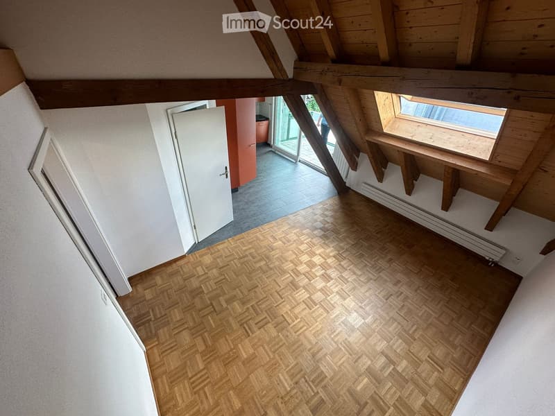 An unfurnished attic room with wooden herringbone flooring, white walls, exposed wooden beams, and a skylight window. The room features a white door leading to another area and a small wooden bench near the window.