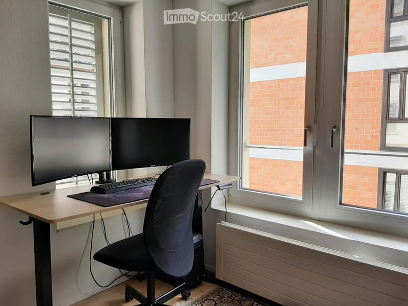A modern, well-lit room with a desk setup featuring a large monitor, keyboard, and mouse, accompanied by a comfortable office chair. The room has large windows with blinds and a radiator below them.