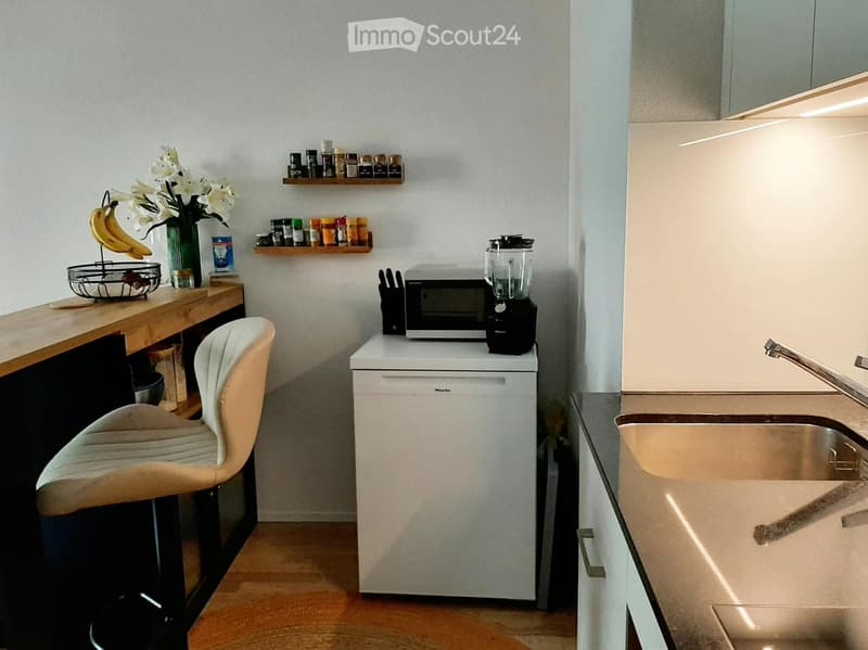 Modern kitchen with white cabinets, a wooden countertop, a stool, a microwave, a dishwasher, a blender, a sink, and decorative shelves with spices and a vase of flowers.