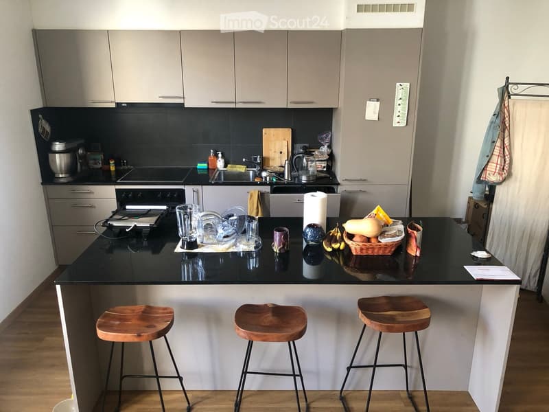 Modern kitchen with grey cabinets, black countertops, and three wooden stools at the island. The island features a blender, cups, a bowl, and a basket of fruit. The background includes an oven, fridge, and sink area.
