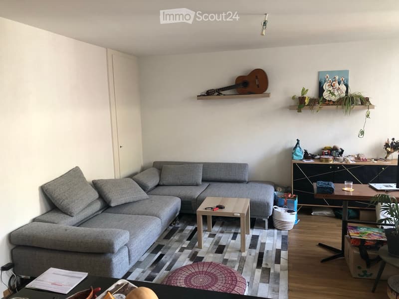 Living room with a grey sectional sofa, wooden coffee table, small round stool, wooden dining table with chairs, guitar on shelf, painting, potted plants, and a cabinet.