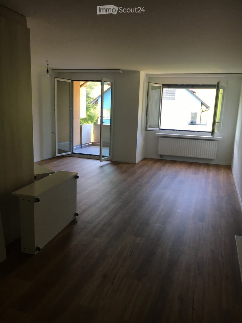 Empty room with wooden flooring, large windows, and an open sliding door leading to a balcony area.