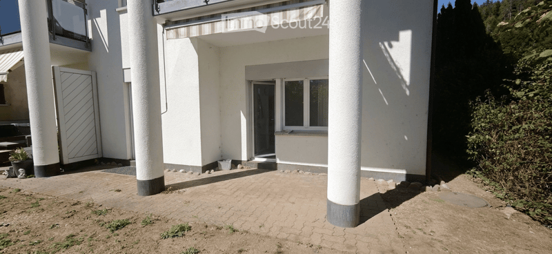 Modern white building with a covered entrance, large windows, and a paved patio area.