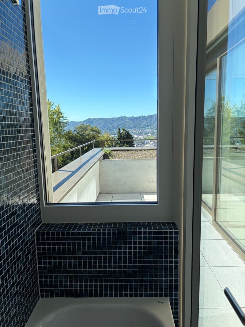Modern bathroom with a bathtub, black tiled walls, and a large window offering a scenic view of the city and mountains.
