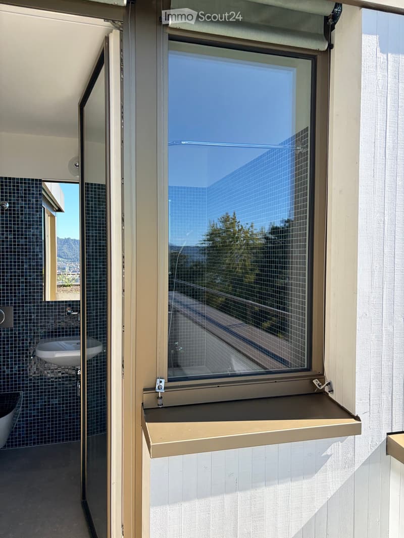 Bathroom with a large window offering a view of a modern building and greenery. The window has a roller shade and a small shelf. The bathroom features a white sink and a dark-tiled bathtub.