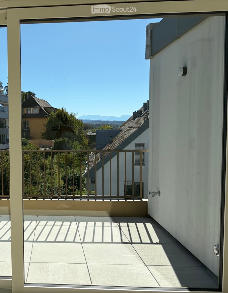 Balcony with tiled floor, metal railing, and a view of surrounding buildings and greenery under a clear blue sky.