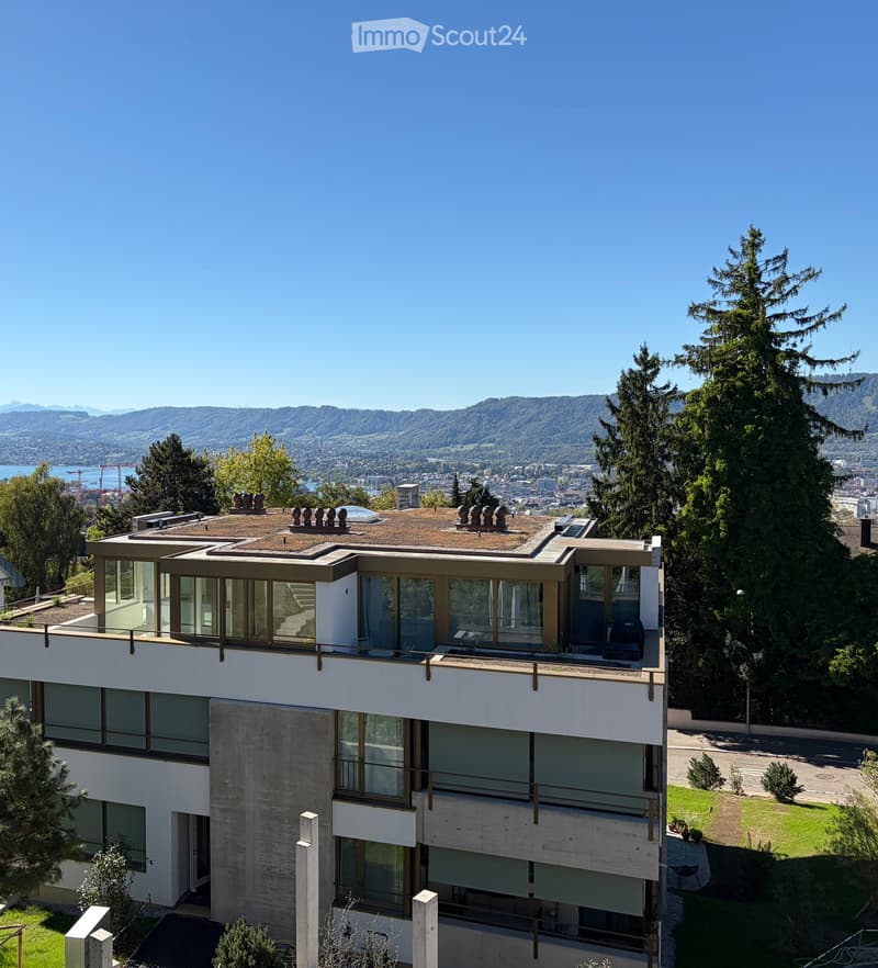 Modern multi-story building with large windows and a flat roof, surrounded by trees and offering a scenic view of a city and mountains.