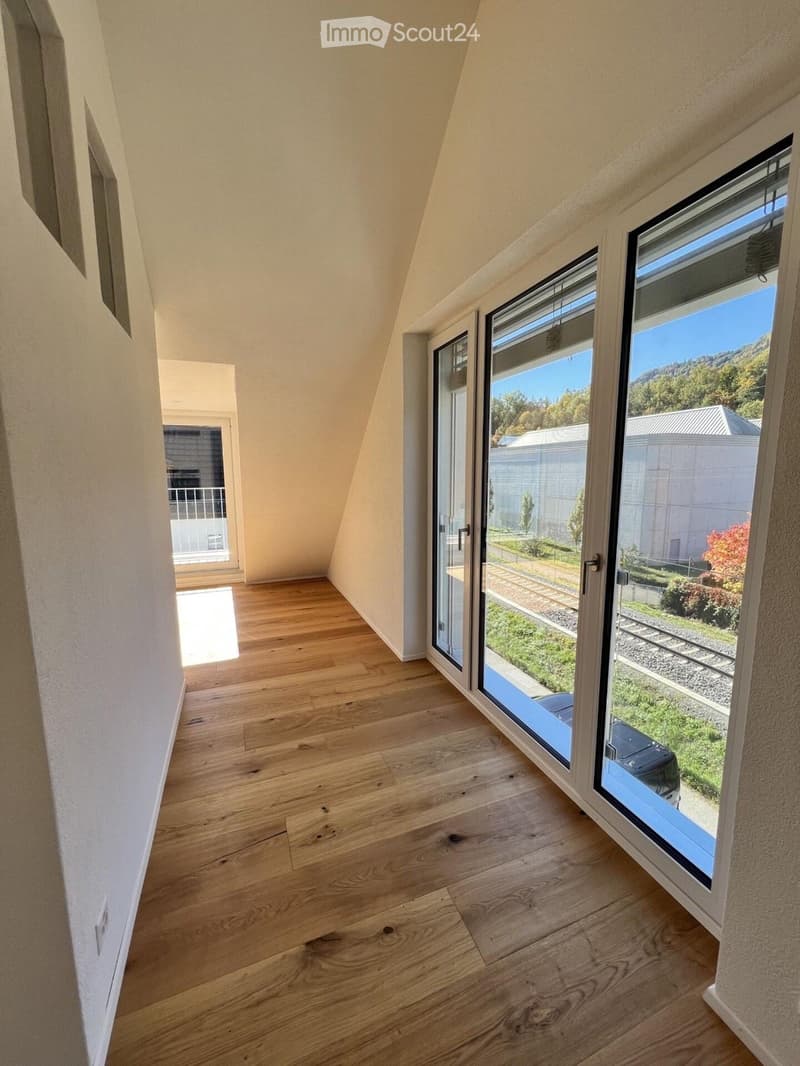 An empty room with wooden flooring and large windows offering a view of the outdoors, including a grassy area and a parked car.