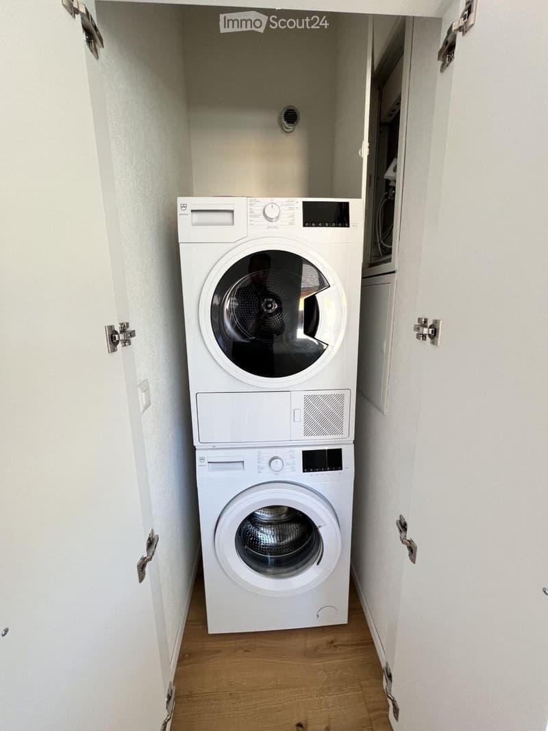 Compact bathroom with a stacked washing machine and dryer unit, white walls, and wooden flooring.