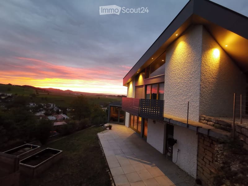Modern two-story house with a balcony, illuminated exterior lighting, and a scenic sunset view over a rural landscape.