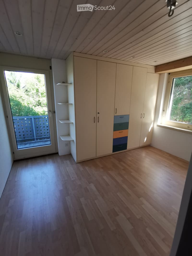 Empty room with light wooden flooring, white walls, built-in wardrobe with multiple drawers and shelves, and a large window with a view of greenery.