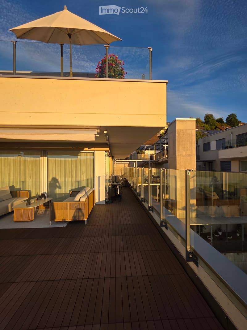 Modern rooftop terrace with wooden decking, glass railing, and outdoor furniture including a sofa, table, and umbrella.