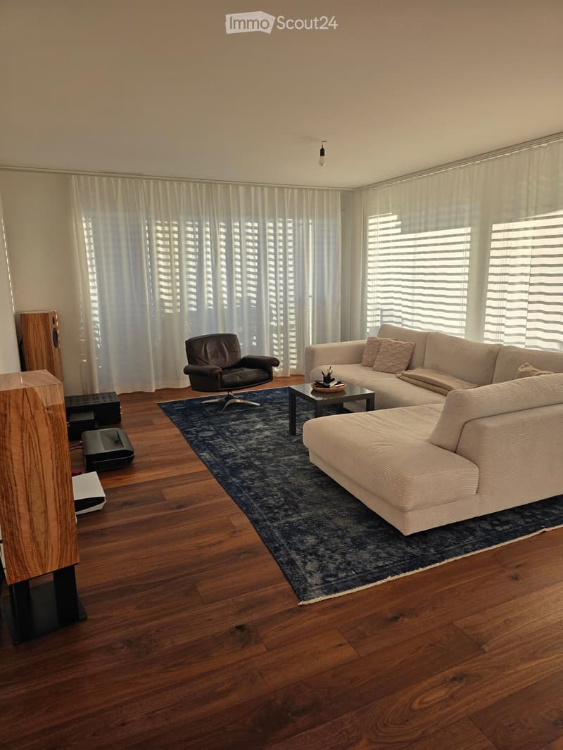Modern living room with wooden flooring, large windows with white curtains, a sectional sofa, armchair, coffee table, speaker system, and a blue rug.