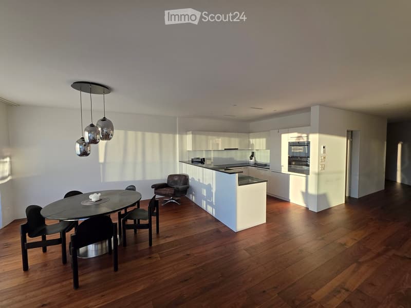 Open-plan dining and kitchen area with wooden flooring, modern white cabinets, a black dining table with chairs, a leather armchair, and pendant lights.