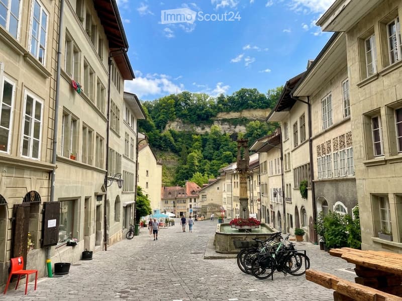 A charming European town square with cobblestone pavement, historic buildings, bicycles, and a small fountain in the center.