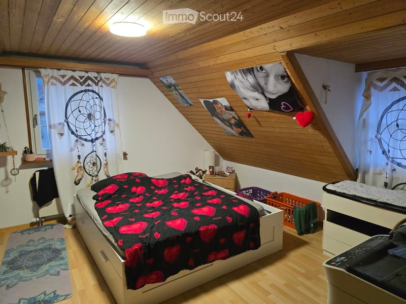 Cozy bedroom with wooden slanted ceilings, a large bed with a black and pink heart-patterned duvet, a nightstand, a dreamcatcher curtain, and decorative items on the walls.