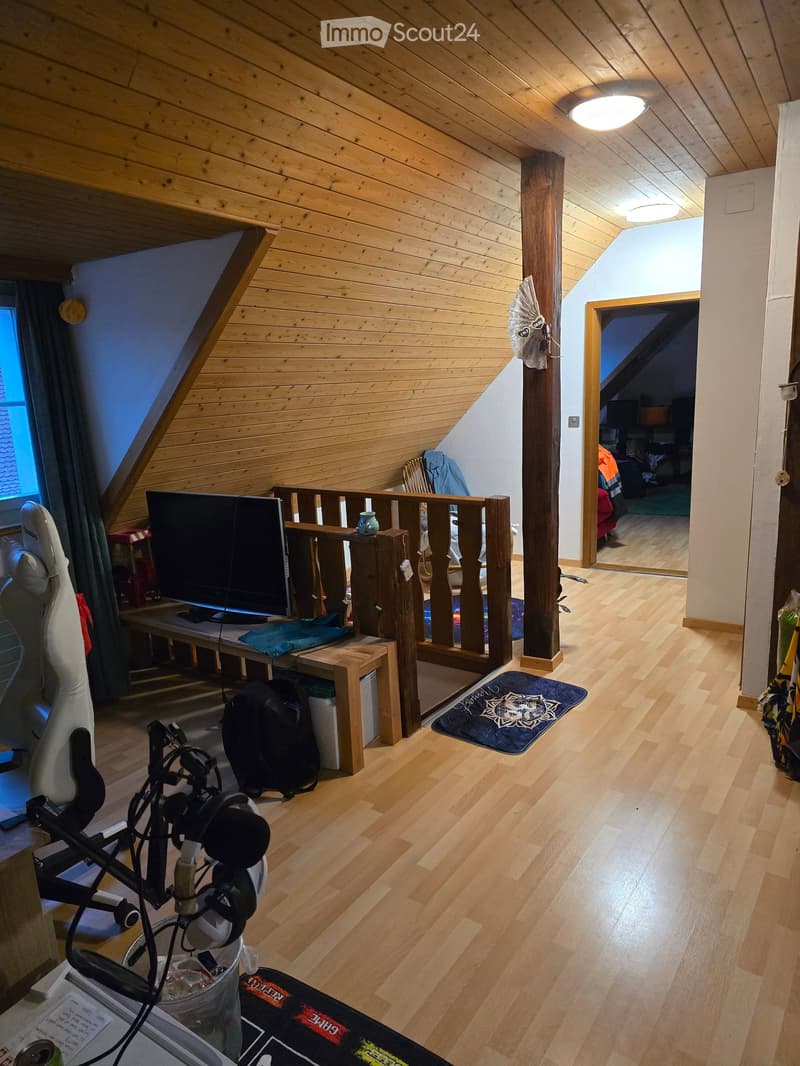 A cozy attic room with wooden walls and ceiling, featuring a table with a screen, a guitar, a backpack, and a camera setup. The room has wooden flooring and a small rug near the doorway.