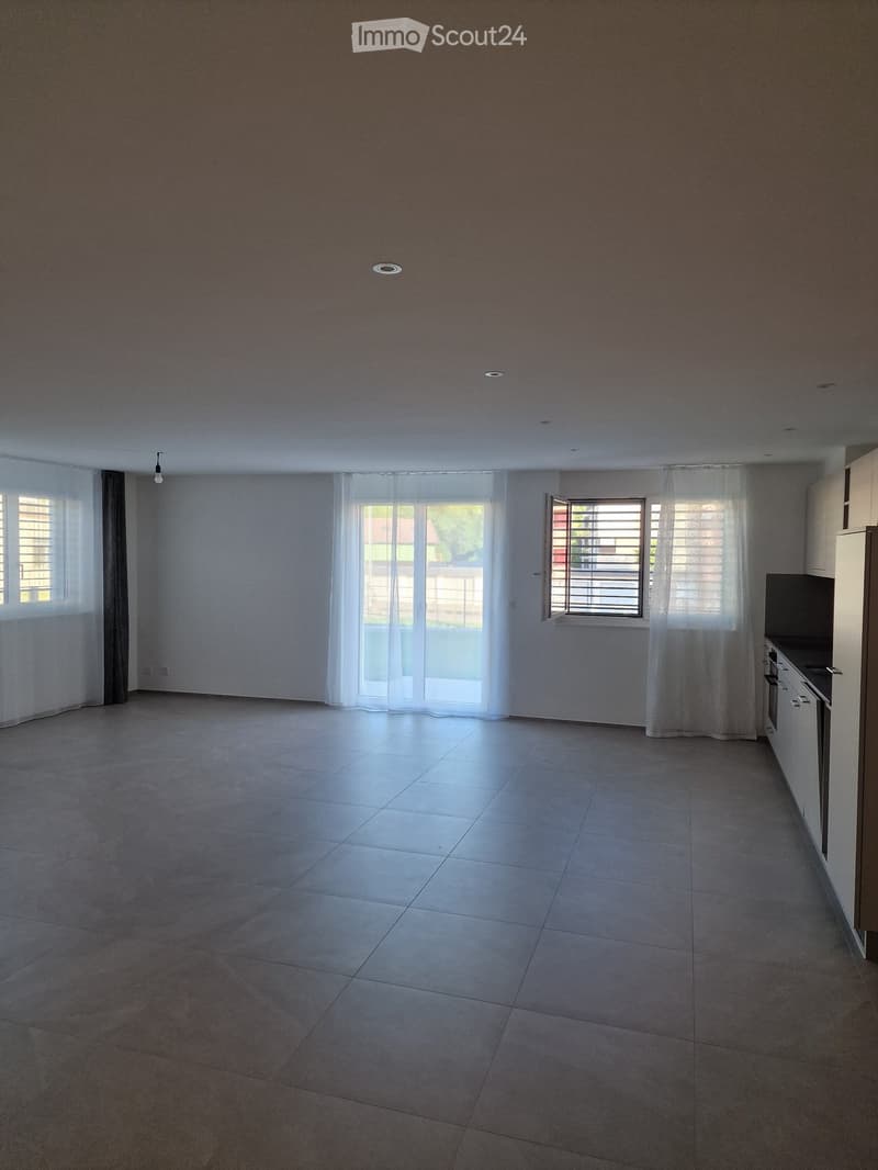 Empty room with tiled floor, white walls, large windows with white curtains, and a small kitchen area with cabinets and appliances visible on the right side.