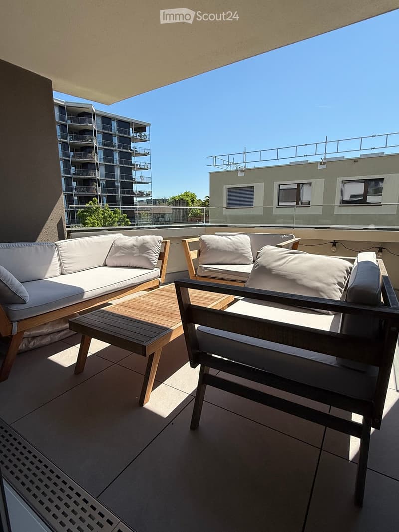 Modern rooftop terrace with a white sofa, wooden chairs, and a small wooden table, overlooking a cityscape with modern buildings.