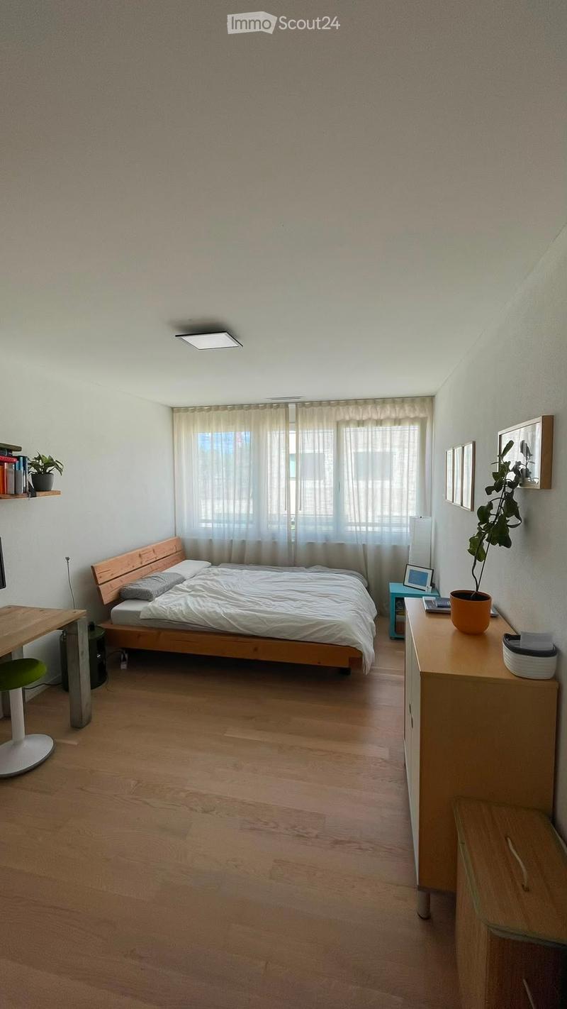 Cozy bedroom with wooden flooring, a bed with white bedding, a nightstand with a lamp, a desk with a chair, and a shelf with books and a plant.