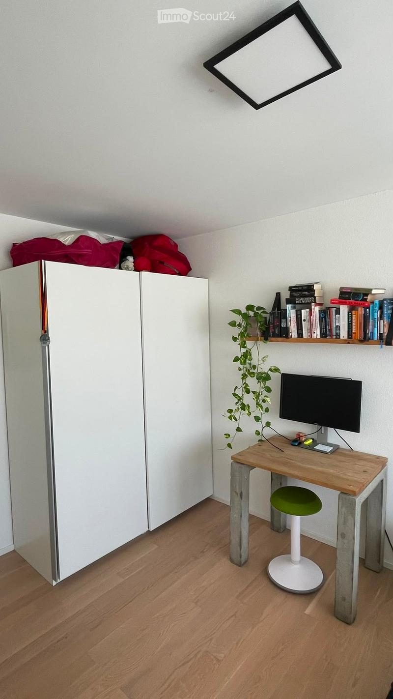 A modern, minimalist room with a wooden floor, a white wardrobe, a wooden desk with a green stool, a computer monitor, a bookshelf with books and a plant, and a square ceiling light.