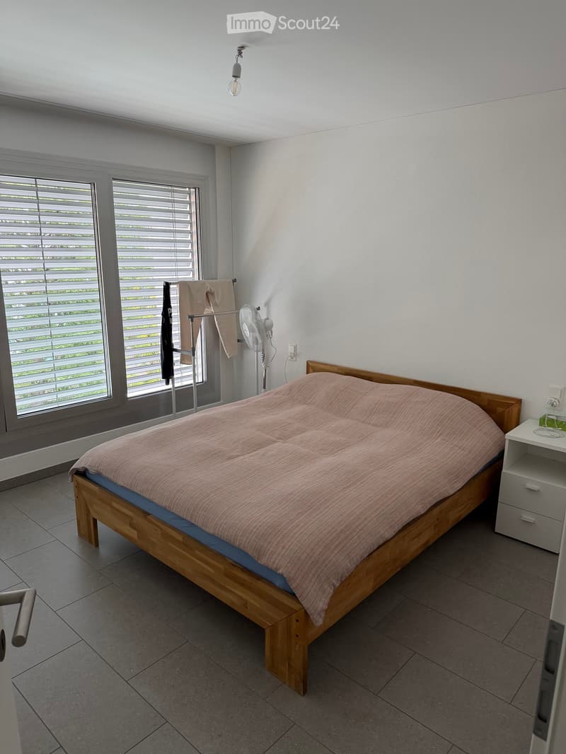 A modern bedroom with a wooden bed, pink bedding, a nightstand, a fan, and a towel rack with towels.
