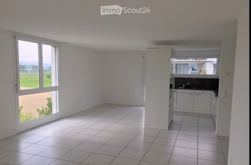 Empty room with white tiled flooring, large windows offering a view of a green field, and a small kitchen area with cabinets and a sink visible in the background.