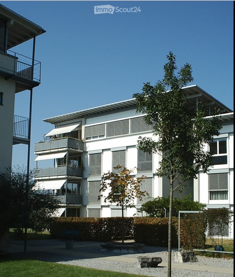 Modern multi-story residential building with white facade, large windows, and balconies. The building features a flat roof and is surrounded by a landscaped area with trees, shrubs, and a stone bench.