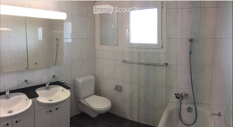 Modern bathroom with beige tiled walls, a double sink vanity, a toilet, a bathtub with a handheld shower, and a large mirror with built-in lighting.