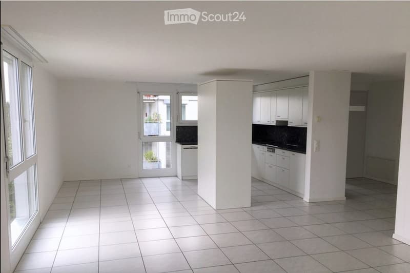 Empty room with white tiled flooring, white walls, and large windows. The space includes a small kitchen area with white cabinets and a countertop.
