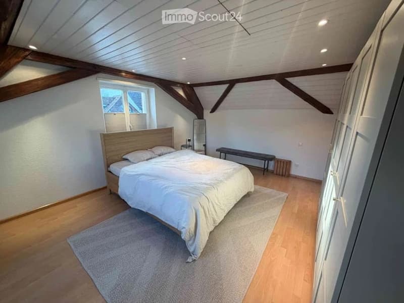 Cozy attic bedroom with wooden beams, white walls, and wooden flooring. Features a large bed with white bedding, a bench, a nightstand, and a mirror.