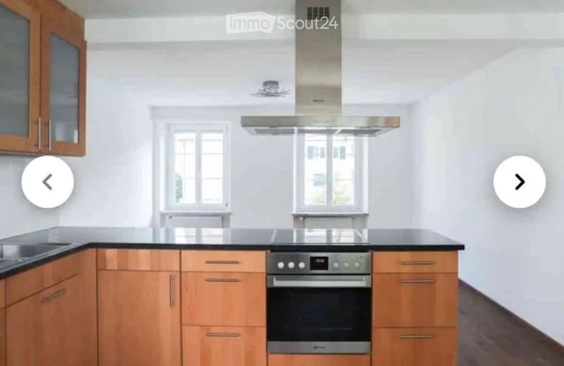 Modern kitchen with light wooden cabinets, black countertops, and stainless steel appliances including a stove and range hood. Two windows provide natural light.