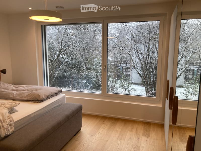 Bright bedroom with large windows overlooking a snowy outdoor scene, featuring a bed with a grey duvet and an ottoman.