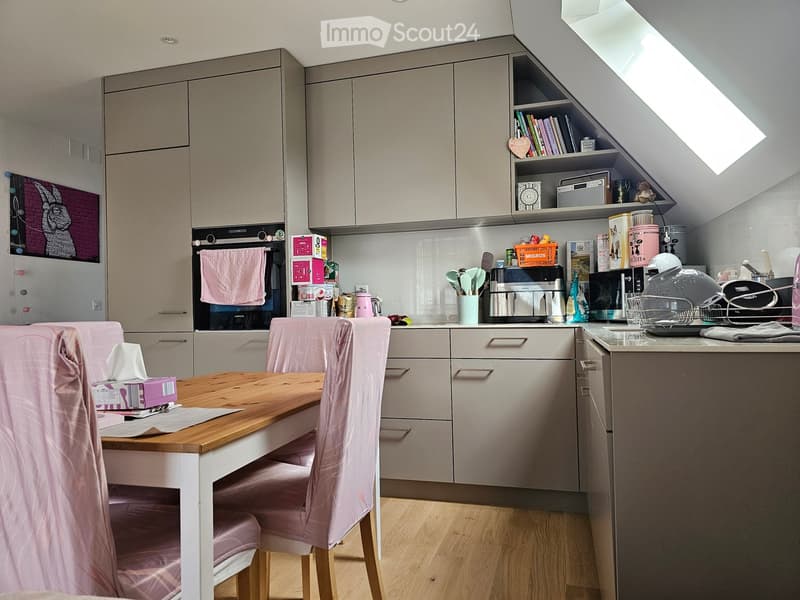 Modern kitchen with grey cabinets, wooden dining table with pink chairs, various appliances including oven, microwave, and fridge, and shelves with books and decorative items.