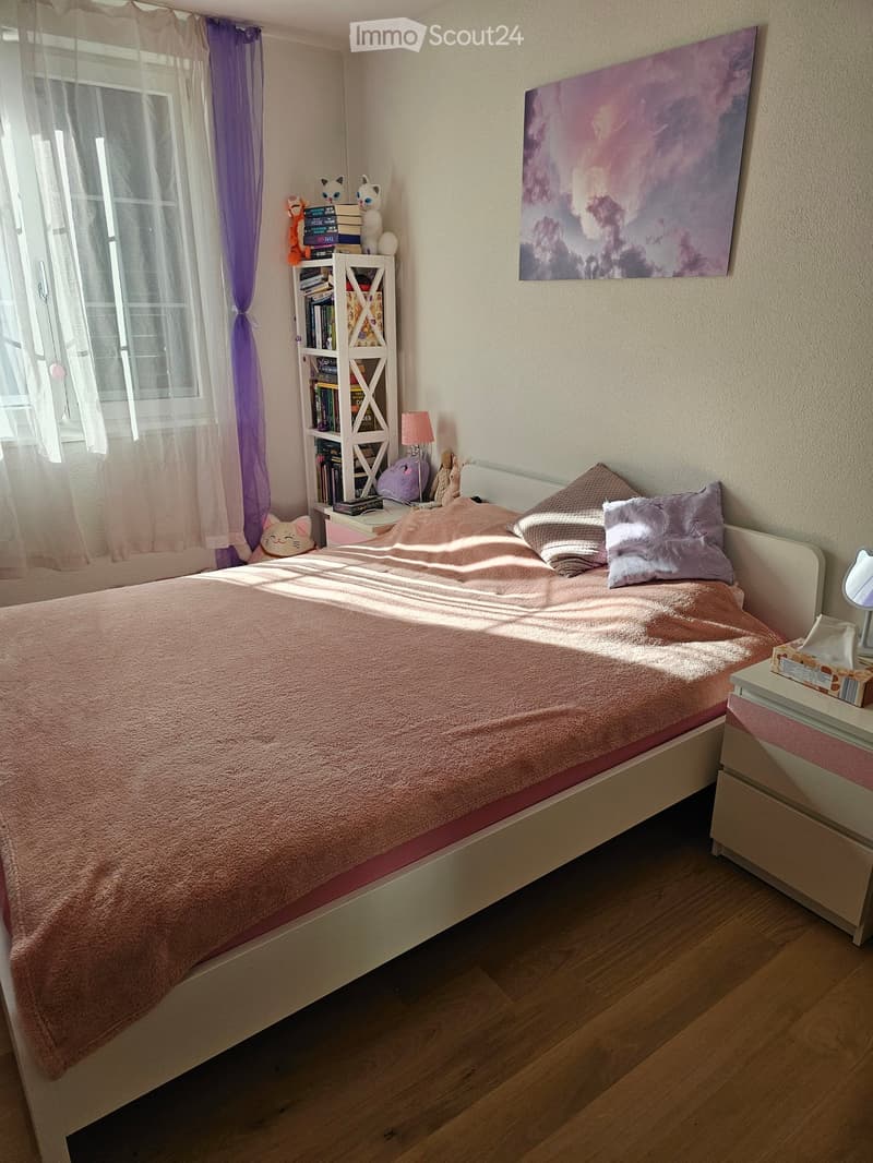 Cozy bedroom with a white bed, pink bedding, nightstand, bookshelf, and decorative items. Natural light streams in through a window with sheer curtains.