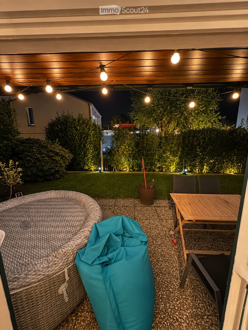 Outdoor patio area with a wooden table, chairs, a wicker hot tub, and string lights overhead. The space is surrounded by greenery and a well-maintained lawn.
