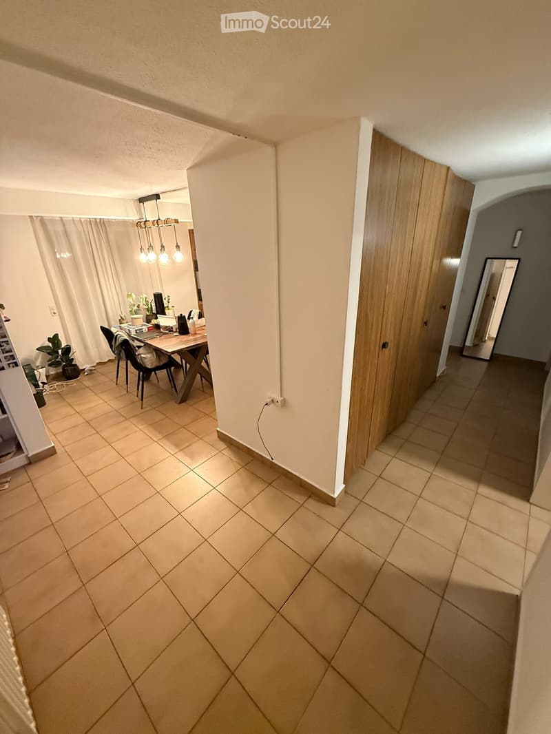 A well-lit dining area with a modern wooden dining table, chairs, a chandelier, and potted plants. The space is connected to a hallway with tiled flooring and built-in wooden cabinets.